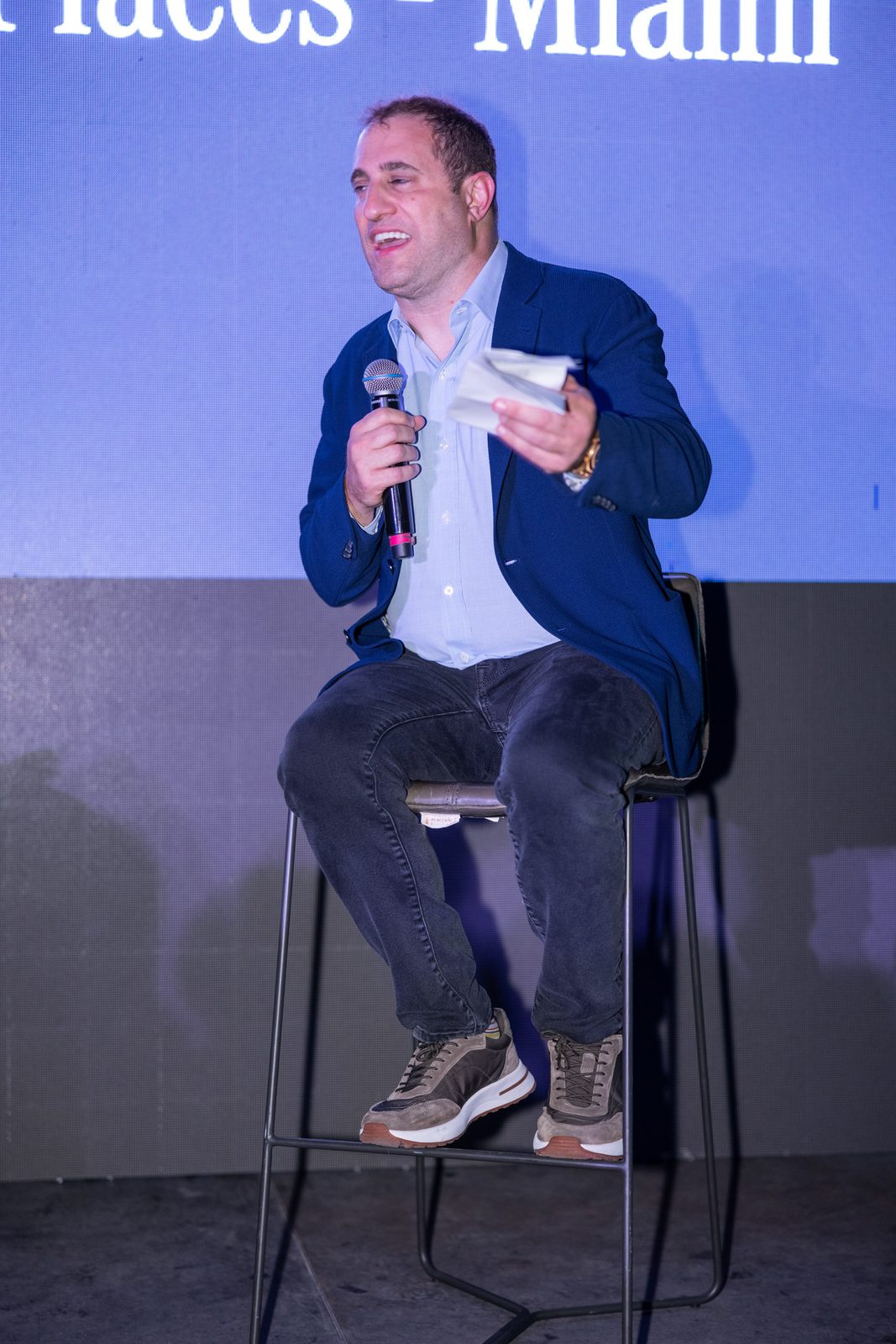 Michael Stern sits on a stool with a microphone in front of a projection screen.